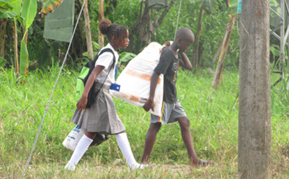 Niño y niña afrodescendientes caminan por una zona verde, él carga un bulto al hombro y ella carga la maleta del colegio.  