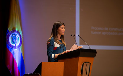Mujer con vestido azul frente a pantalla de auditorio, habla en atril un discurso.