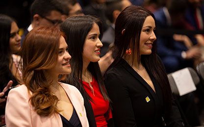 Tres mujeres sentadas, sonríen durante ceremonia de grados Uniandes.