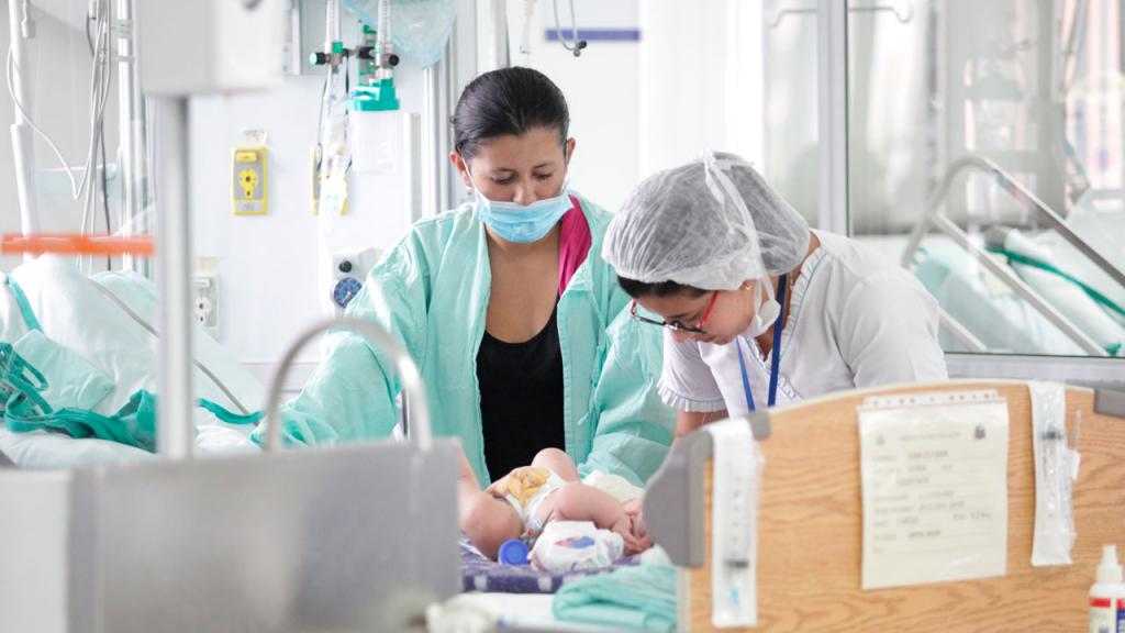 Una enfermera atiende a un bebe en un hospital. La madre observa. 