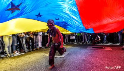 Protestas en Venezuela, un joven corre bajo una bandera venezolana.